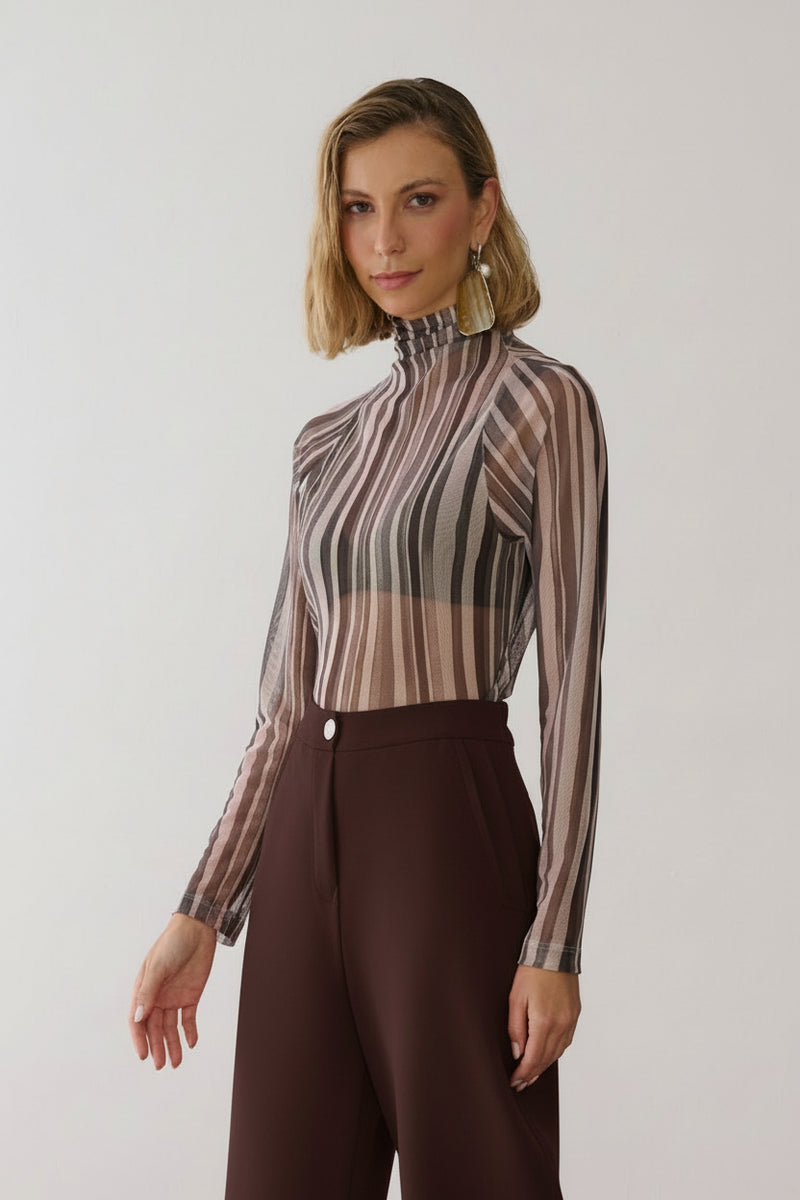 Modelo veste uma blusa de gola alta e mangas longas em tule com estampa de listras verticais em tons de off e cinza, combinada com calça de alfaiataria marrom.