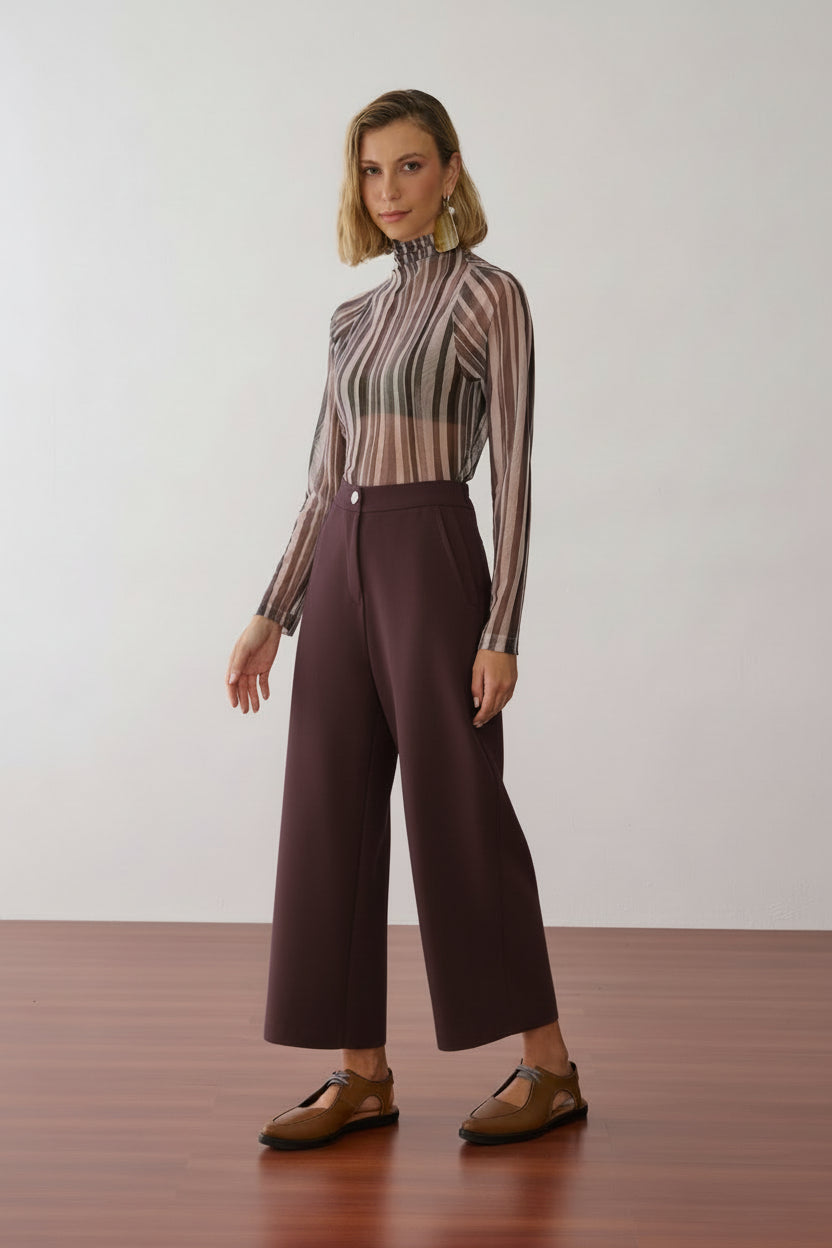 Modelo veste uma blusa de gola alta e mangas longas em tule com estampa de listras verticais em tons de off e cinza, combinada com calças pantalona marrom e sapatos marrons de bico fino com recortes laterais.