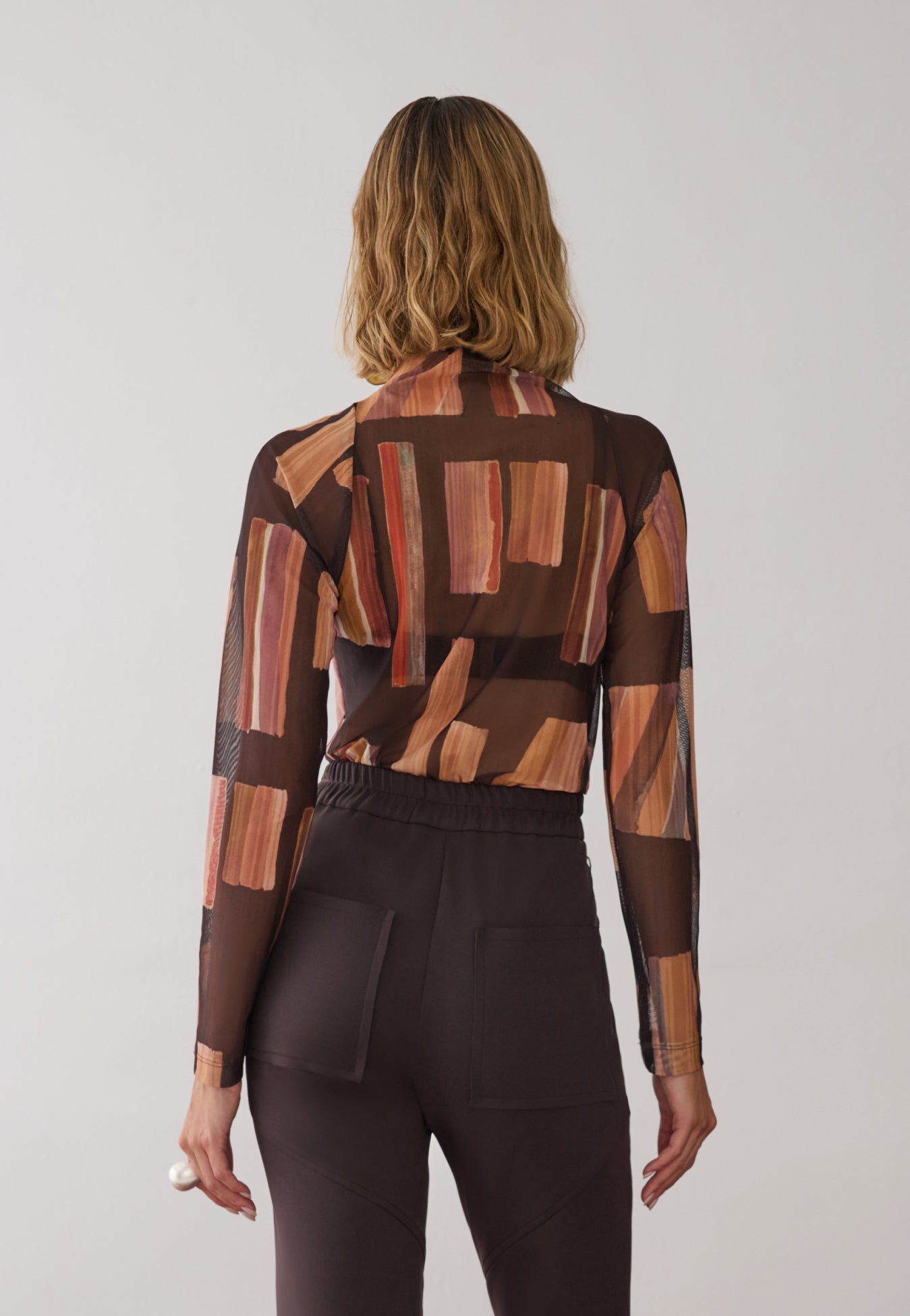 Modelo de costas veste uma blusa de gola alta e mangas longas em tule com estampa geométricas em tons de marrom, combinada com calça de alfaiataria marrom de cós elástico e bolsos amplos.