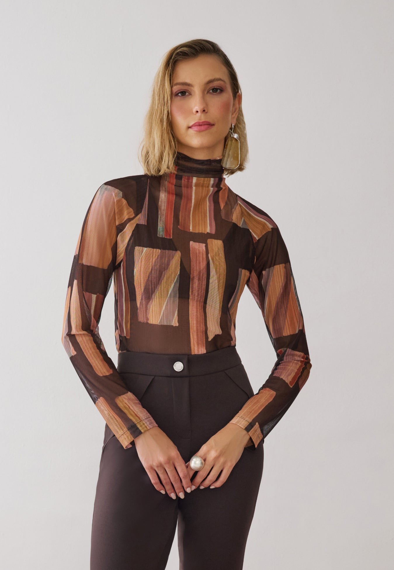 Modelo veste uma blusa de gola alta e mangas longas em tule com estampa geométricas em tons de marrom, combinada com calças de alfaiataria marrom.