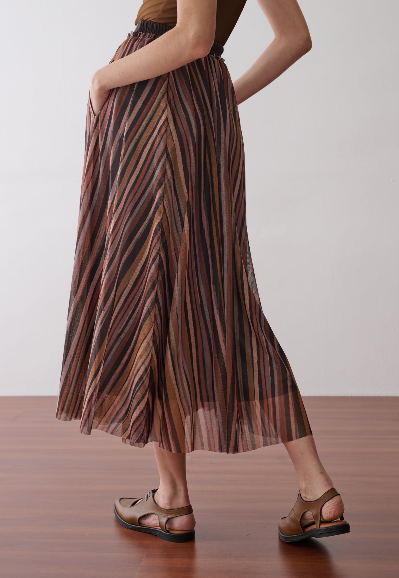 Modelo de costas veste uma saia longa plissada em tule com estampa de listras multicoloridas.