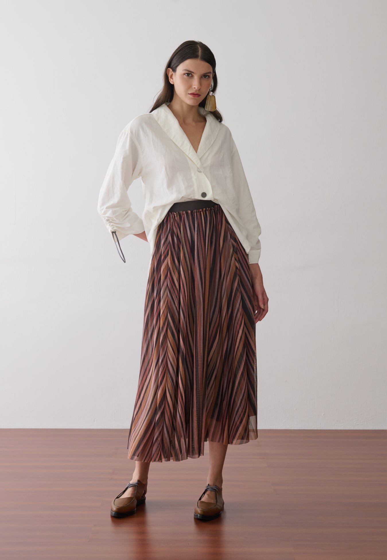 Modelo veste uma camisa off-white com gola "V" e mangas longas com ajuste na barra, combinada com uma saia longa plissada em tule com estampa de listras multicoloridas.