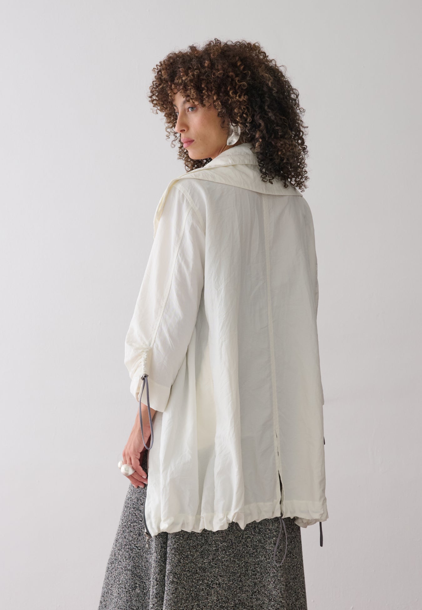 Modelo de costas usando uma jaqueta off-white de tecido leve com ajuste por cordões e reguladores na barra e mangas, sobre um vestido mescla cinza de caimento amplo.