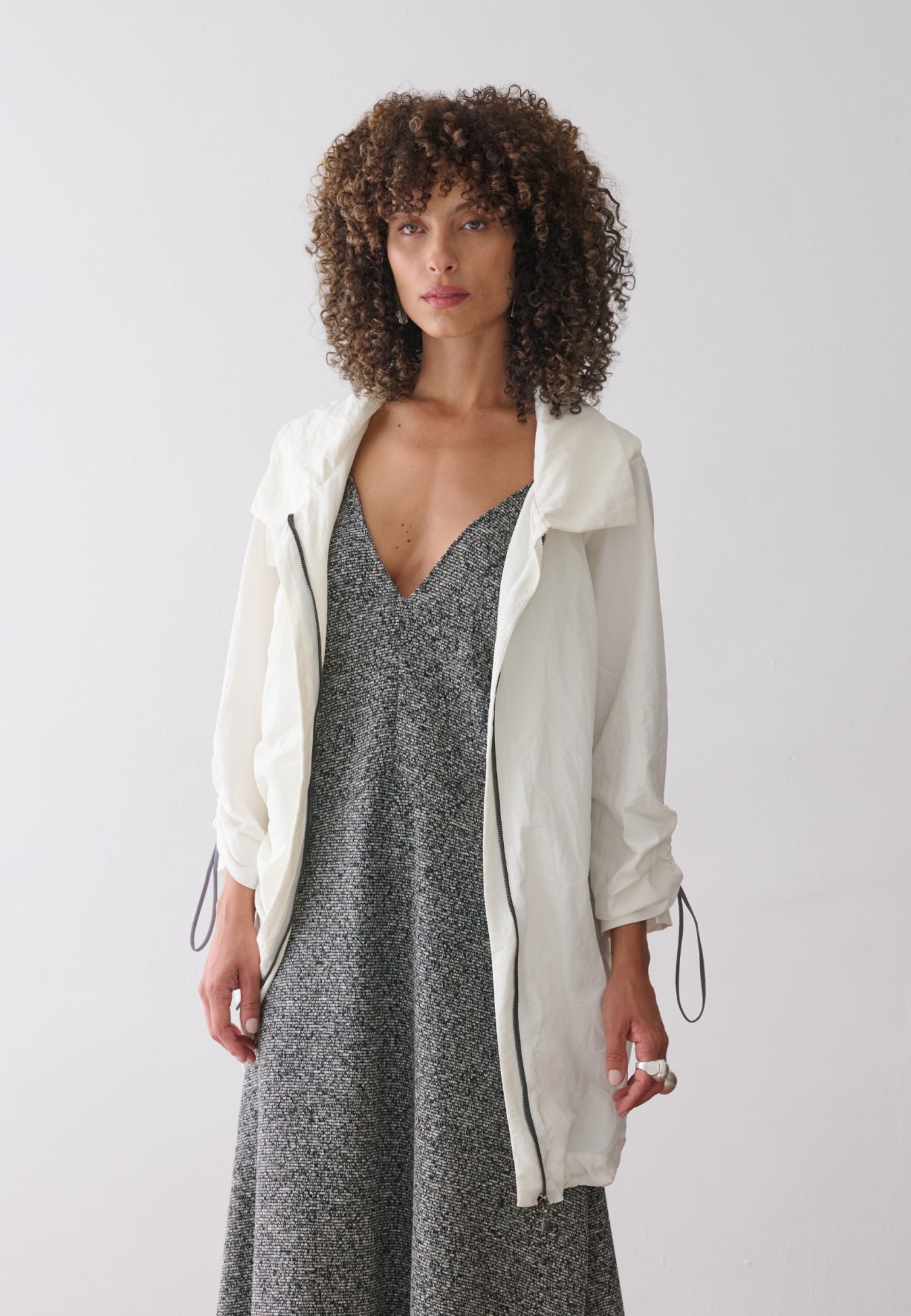 Modelo veste uma jaqueta longa off-white de tecido leve, mangas ajustadas com cordões e reguladores, sobre um vestido mescla cinza de decote em "V".