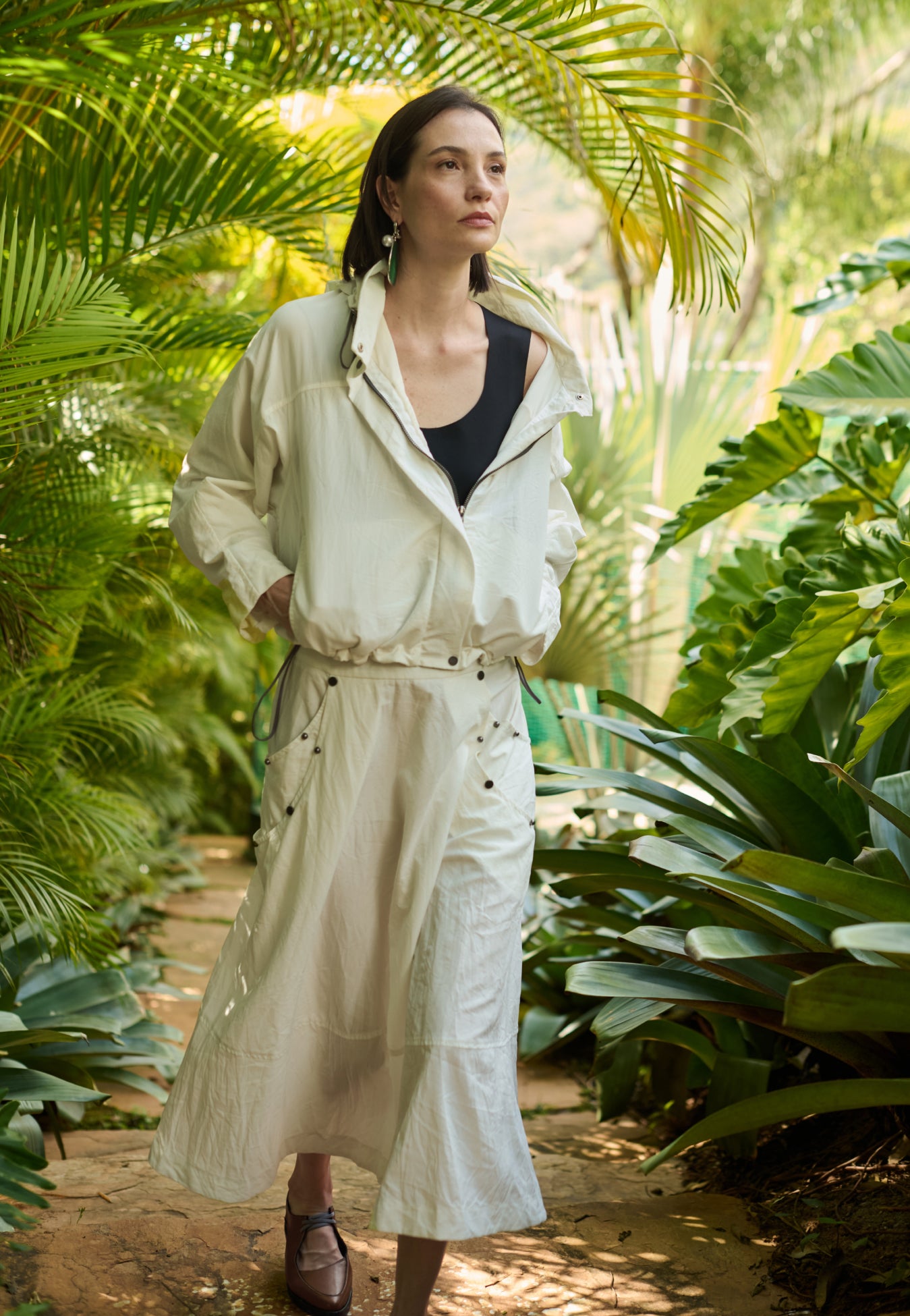 Modelo veste uma jaqueta off-white com capuz sobre uma regata preta, combinada com uma saia midi off-white que possui bolsos laterais utilitários com rebites e sapatos marrons de bico fino.