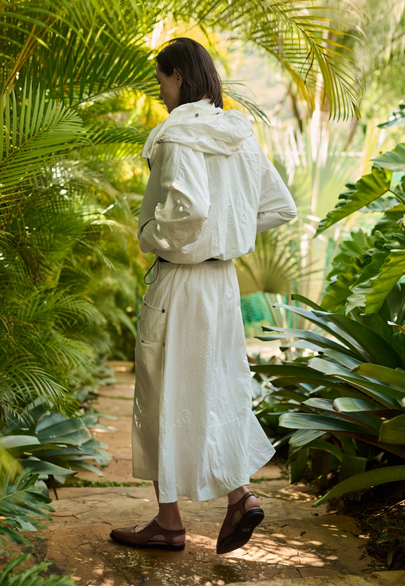 Modelo de costas veste uma jaqueta off-white com capuz, combinada com uma saia midi off-white que possui bolsos laterais utilitários com rebites e sapatos marrons de bico fino.