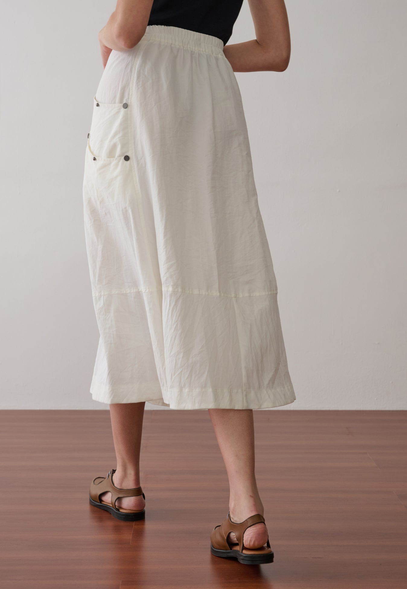 Modelo de costas veste uma saia midi off-white, possui cós elástico, recorte horizontal na barra, bolsos laterais utilitários com rebites e sapatos marrons de bico fino.