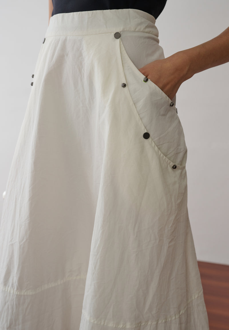 Modelo veste uma saia midi off-white que possui bolsos laterais utilitários com rebites e aplicações.