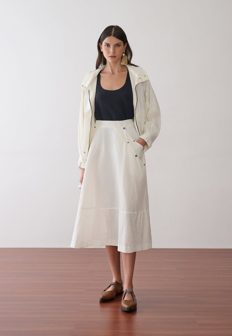 Modelo veste uma jaqueta off-white com capuz sobre uma regata preta, combinada com uma saia midi off-white que possui bolsos laterais utilitários com rebites e sapatos marrons de bico fino.