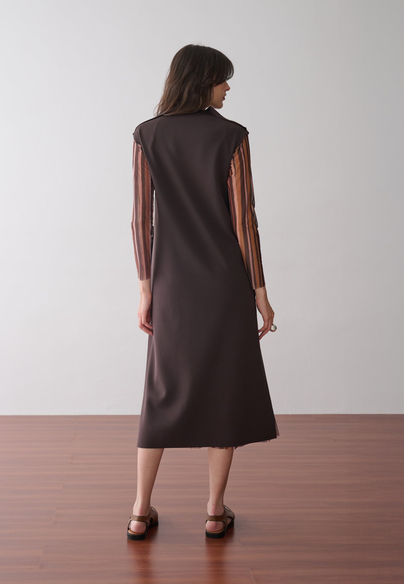 Modelo de costas usa um vestido midi marrom de gola alta com forro removível em tule de estampa listrada multicolorida, combinados com sapatos marrons de bico fino.