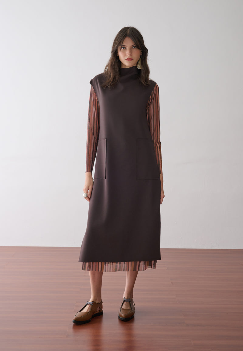 Modelo usa um vestido midi marrom de gola alta e bolsos frontais com  forro removível em tule de estampa listrada multicolorida, combinados com sapatos marrons de bico fino.