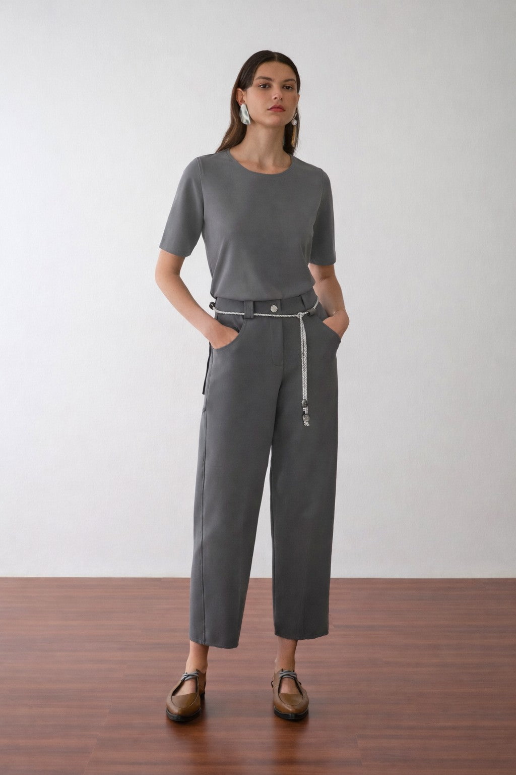 Modelo veste camiseta cinza de mangas curtas, combinada com calça de alfaiataria na mesma cor, cordão off white na cintura e sapatos marrons de bico fino.