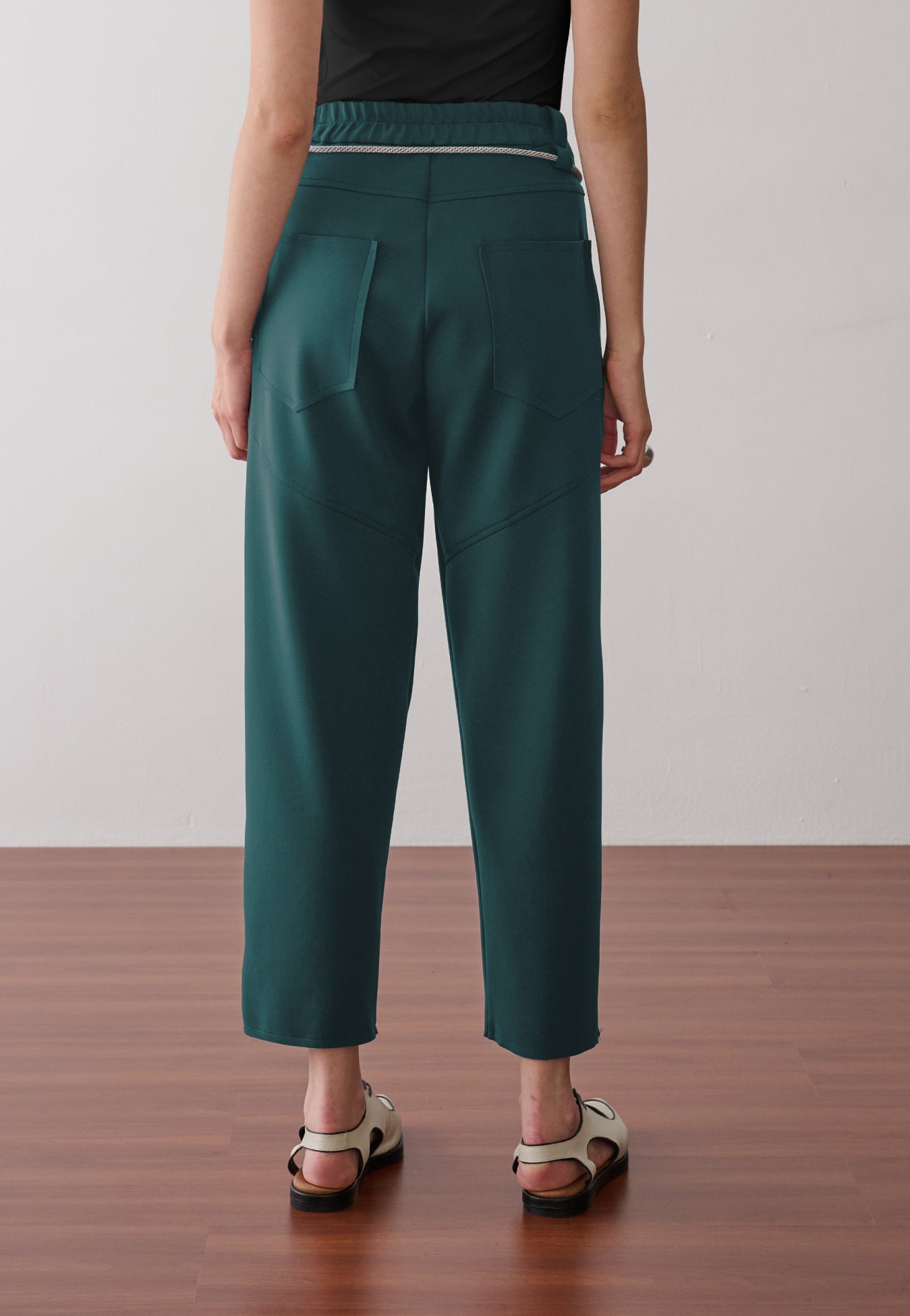 Modelo de costas usando uma calça verde-petróleo de cintura alta com cós elástico, bolsos traseiros amplos com costuras geométricas e sandálias off white.