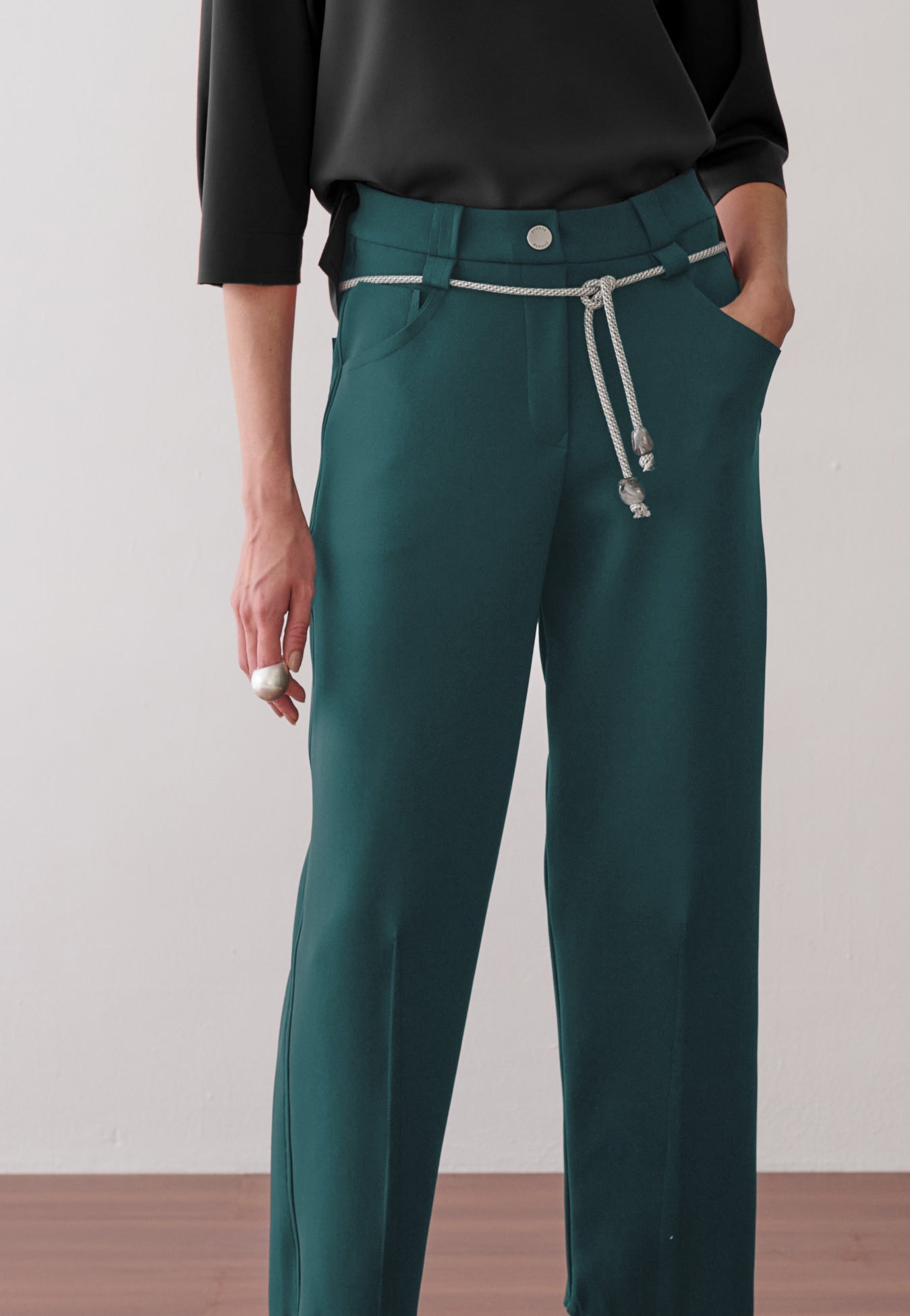 Modelo veste calça de alfaiataria verde-petróleo com vincos frontais, acompanhada de um cordão prateado com ponteiras na cintura.