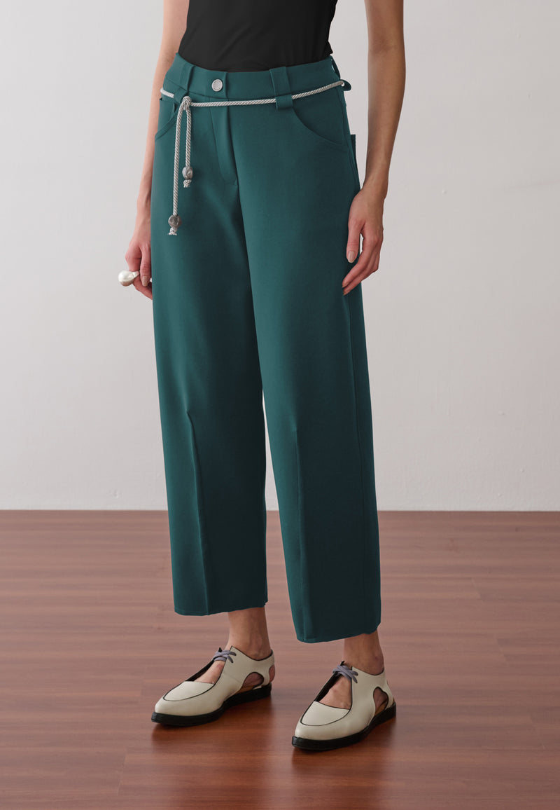 Modelo veste calça de alfaiataria verde-petróleo com vincos frontais, acompanhada de um cordão prateado com ponteiras na cintura e sapatos off-white de bico fino.