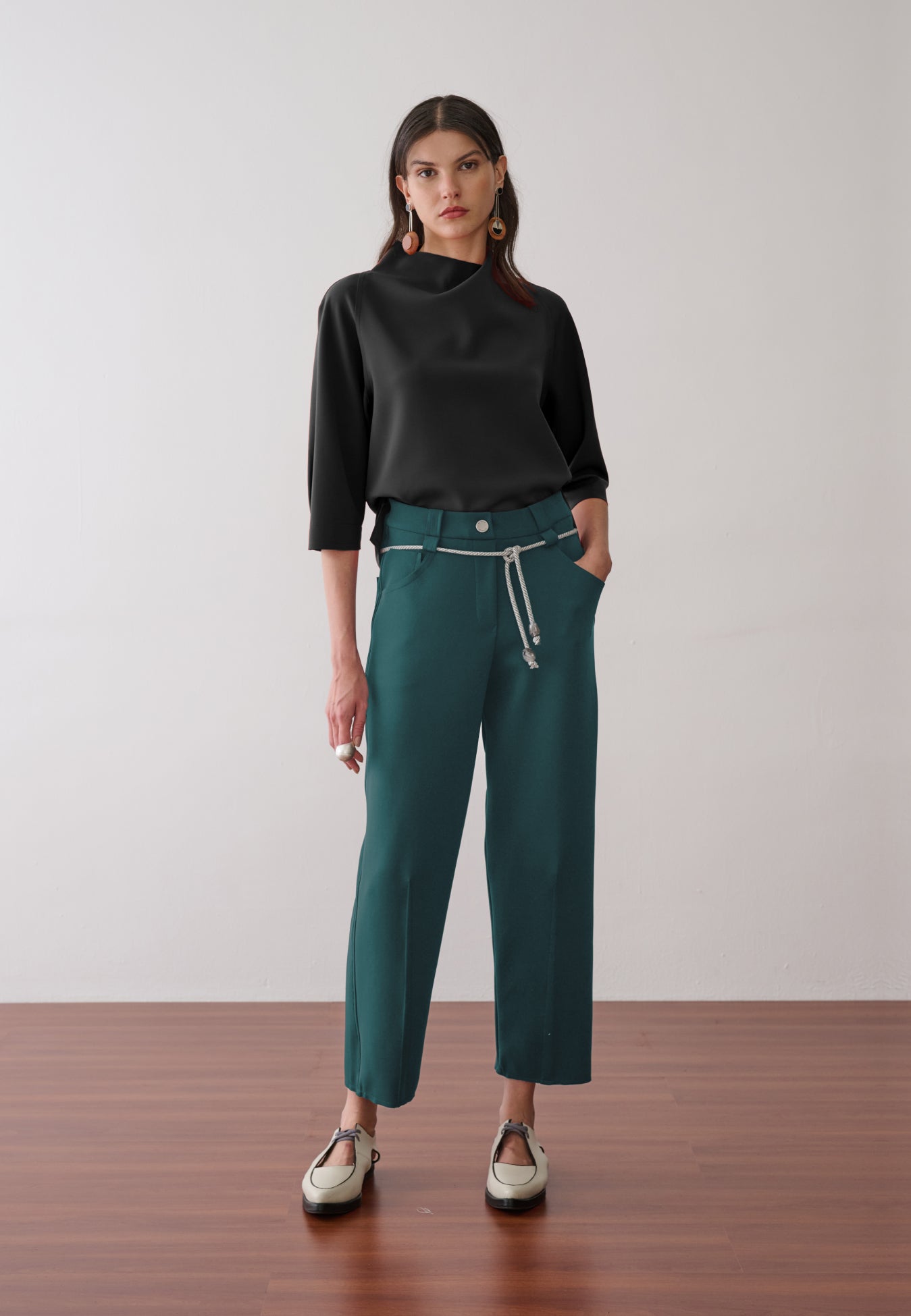 Modelo veste uma blusa preta de gola alta e mangas 3/4, combinada com calça de alfaiataria verde-petróleo, cordão prateado na cintura e sapatos off-white de bico fino com recortes.