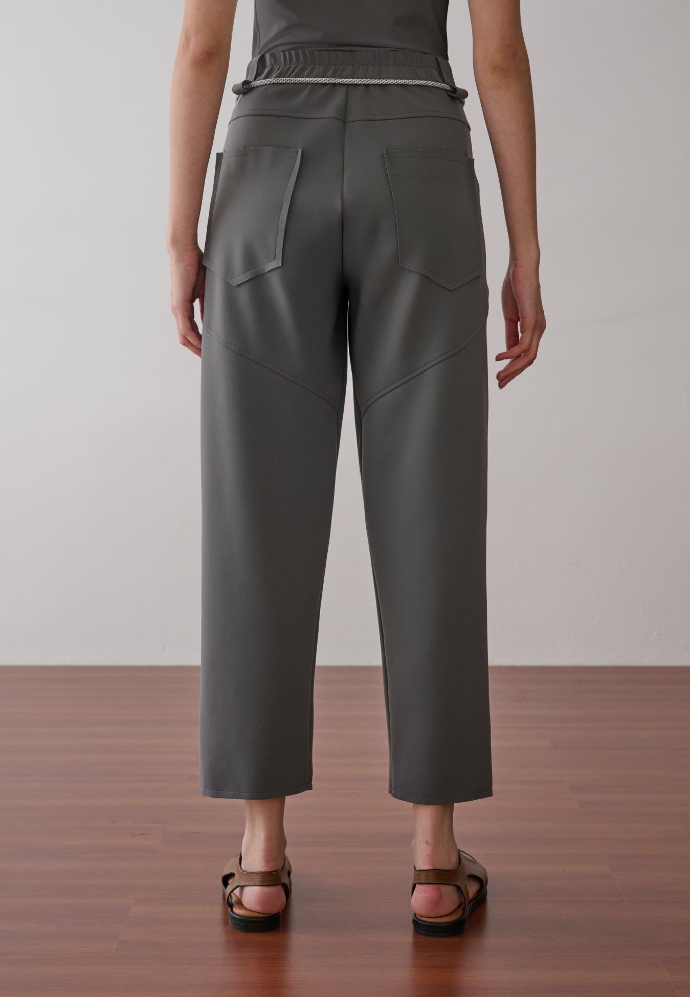 Modelo de costas usando uma calça cinza de cintura alta com cós elástico, bolsos traseiros amplos com costuras geométricas e sandálias marrons.