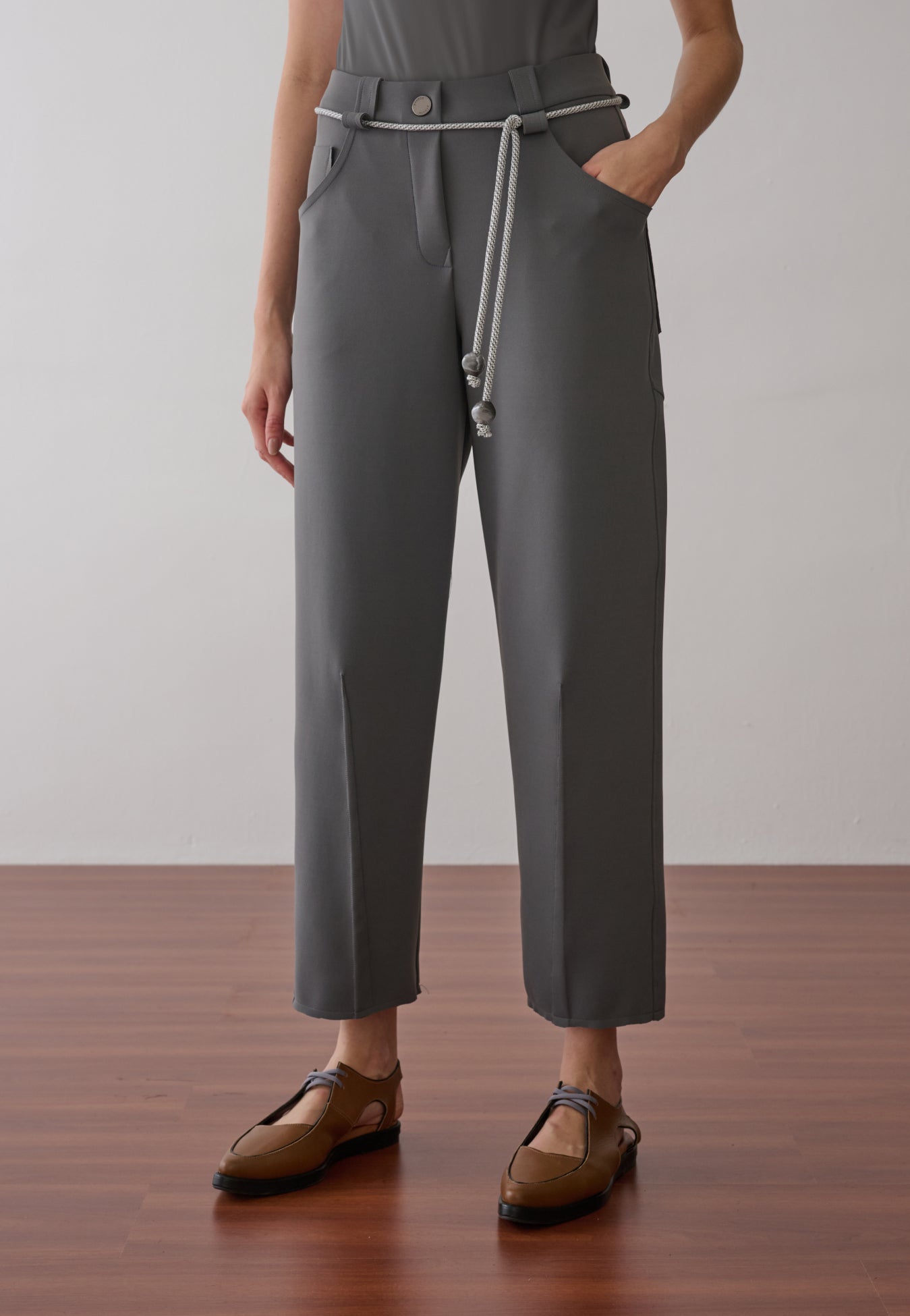 Modelo veste calça de alfaiataria cinza com vincos frontais, acompanhada de um cordão prateado com ponteiras na cintura e sapatos marrons de bico fino.