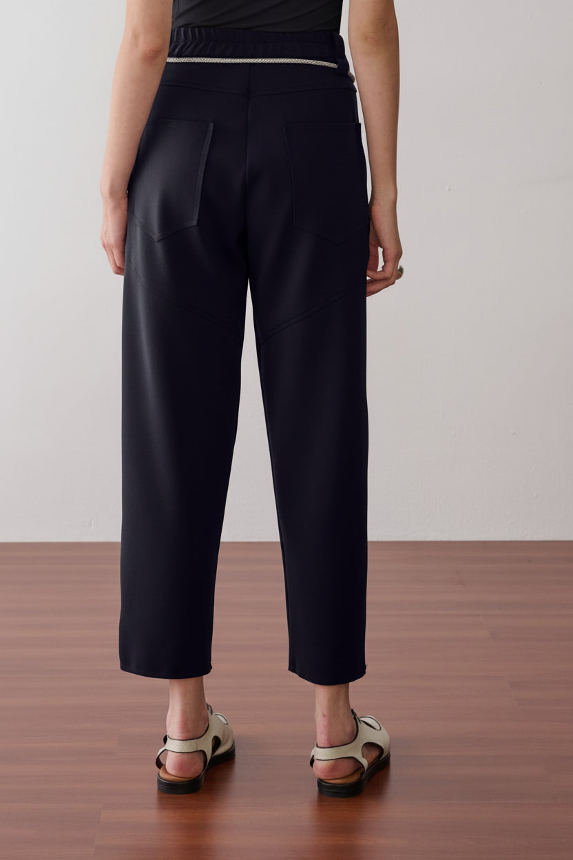 Modelo de costas usando uma calça preta de cintura alta com cós elástico, bolsos traseiros amplos com costuras geométricas e sandálias off white.