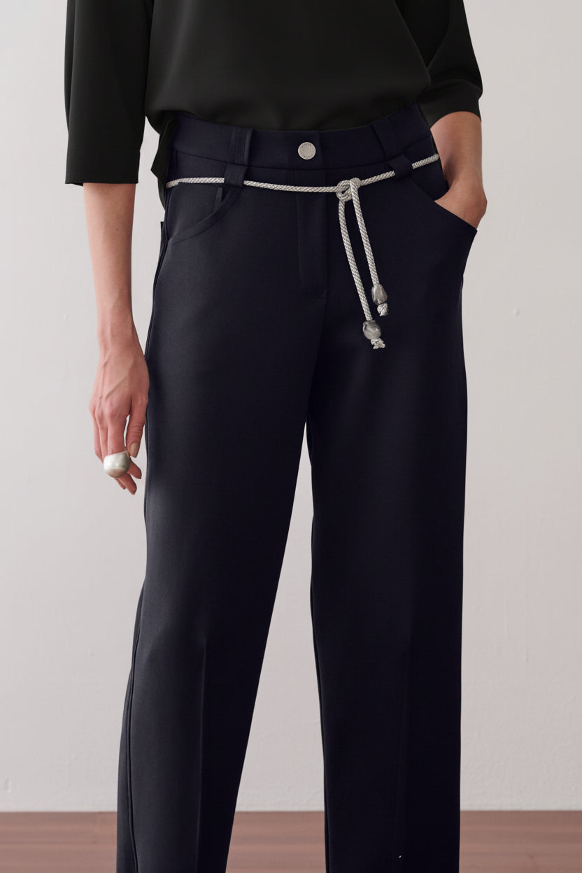 Modelo veste calça de alfaiataria preto com vincos frontais, acompanhada de um cordão prateado com ponteiras na cintura.