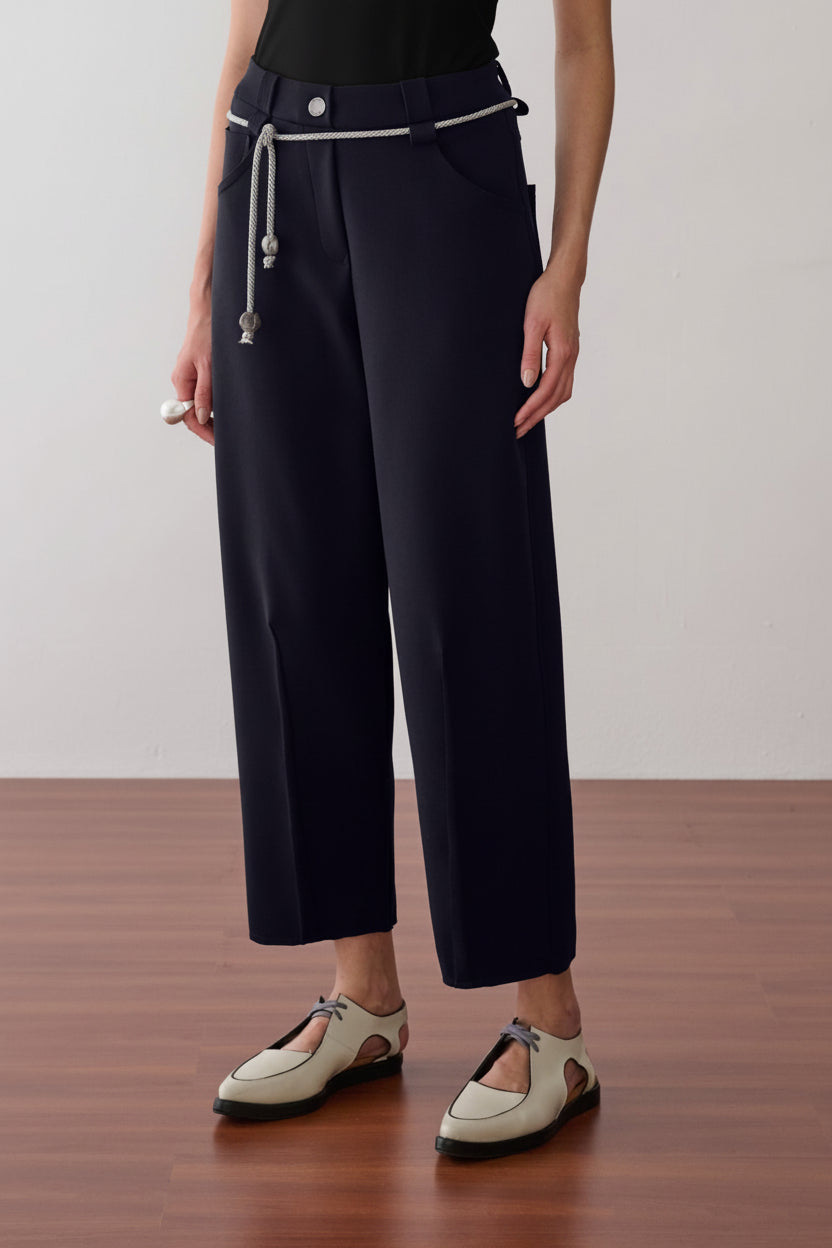 Modelo veste calça de alfaiataria preta com vincos frontais, acompanhada de um cordão prateado com ponteiras na cintura e sapatos off-white de bico fino.