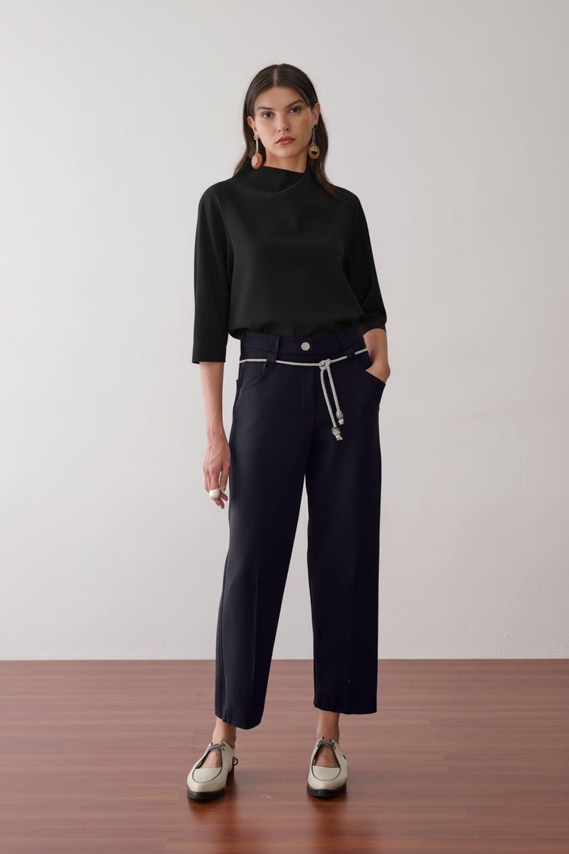 Modelo veste uma blusa preta de gola alta e mangas 3/4, combinada com calça da mesma cor, cordão prateado na cintura e sapatos off-white de bico fino com recortes.