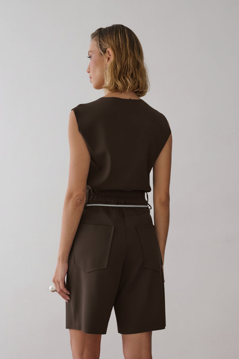 Modelo de costas usando uma blusa marrom sem mangas e bermuda de alfaiataria na mesma cor com bolsos traseiros amplos, detalhe de cós elástico e um cinto de cordão off white.