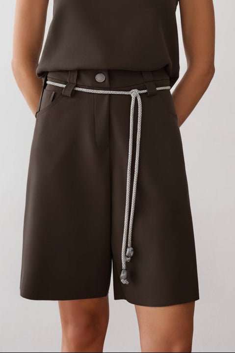 Modelo veste uma bermuda de alfaiataria marrom com botão metalizado, passantes e cordão off white na cintura.