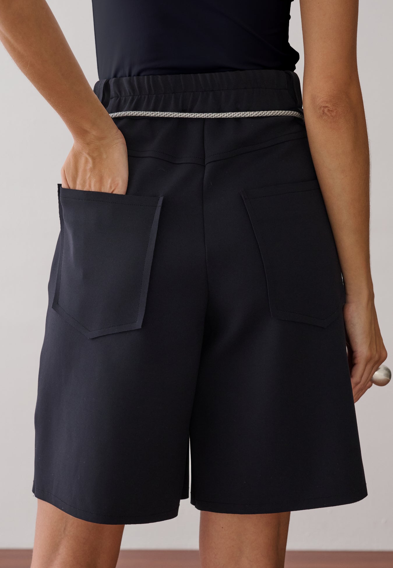Modelo de costas usando uma bermuda de alfaiataria preta com bolsos traseiros amplos, detalhe de cós elástico e um cinto de cordão off.