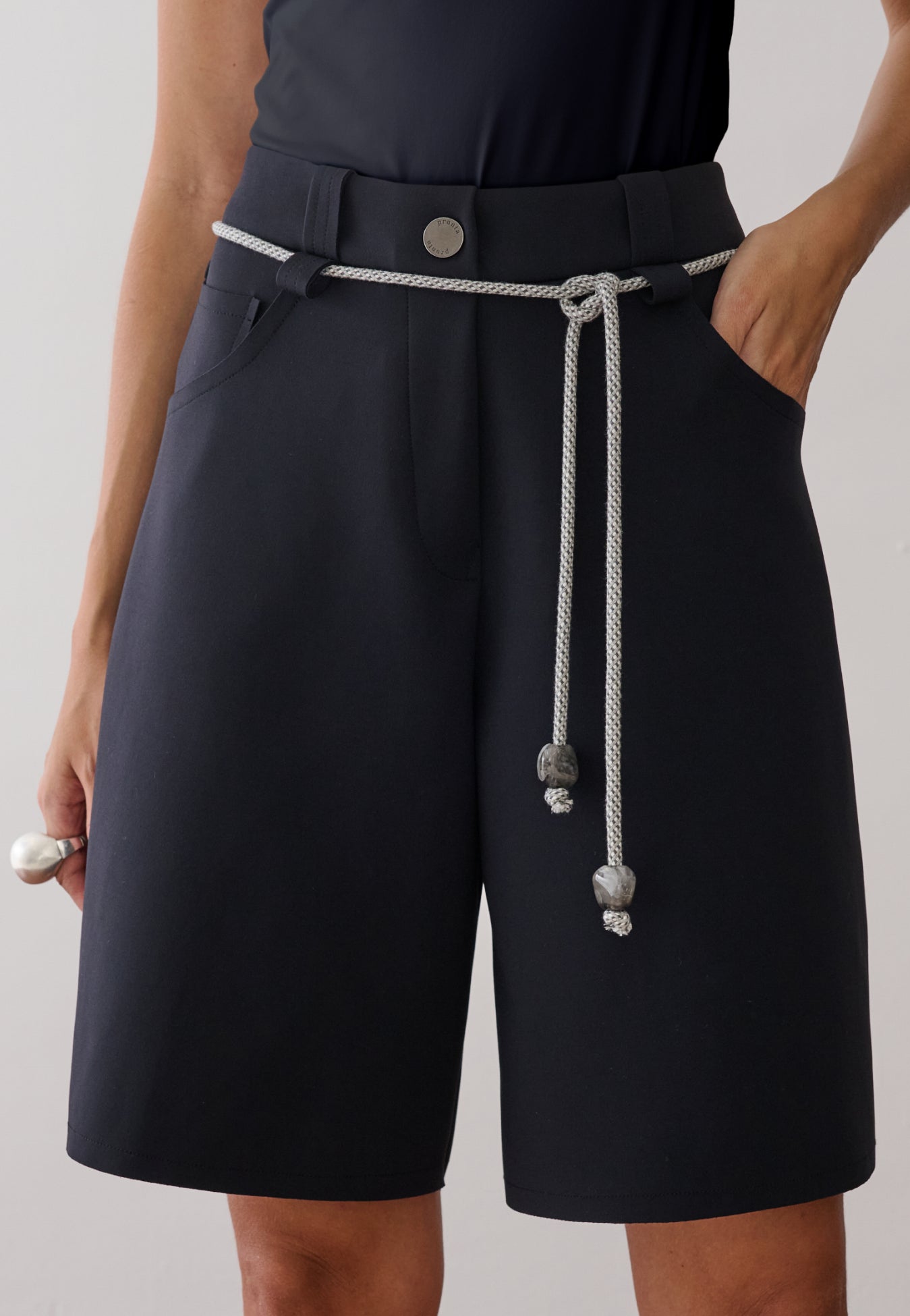 Modelo veste uma bermuda de alfaiataria preta com botão metalizado, passantes e cordão off white na cintura.