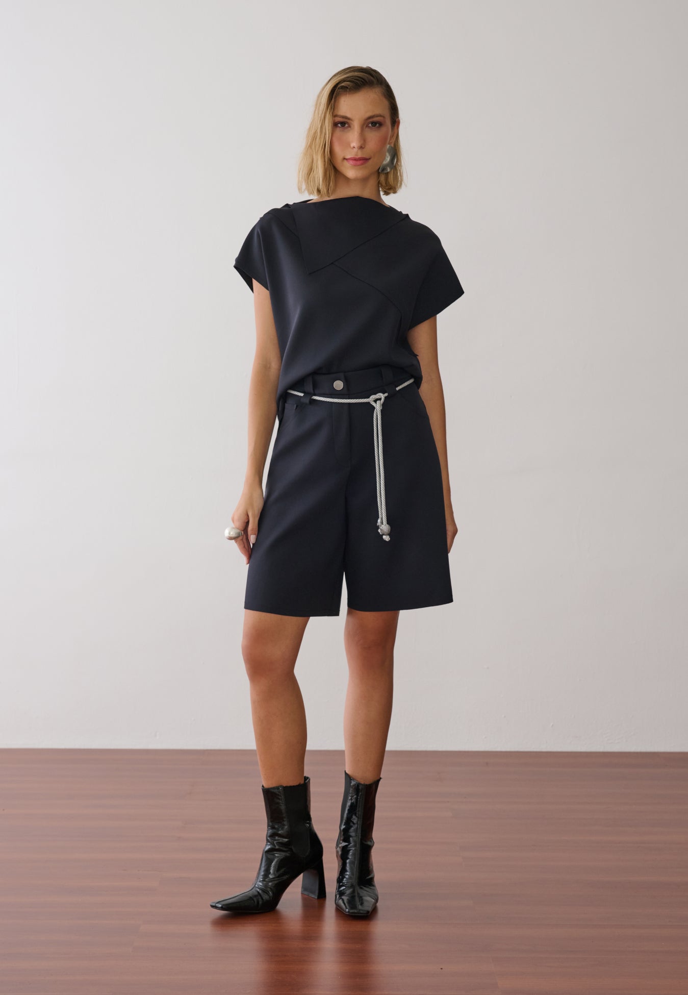 Modelo veste uma blusa preta de gola alta e mangas curtas com costuras geométricas, combinada com bermuda de alfaiataria na mesma cor, cordão off white na cintura e botas pretas de verniz.
