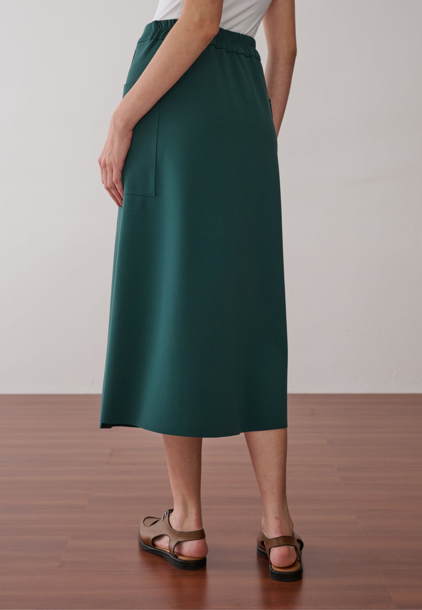 Modelo de costas usando uma saia midi verde-petróleo de acabamento acetinado com cós elástico e bolsos laterais, combinada com blusa de tule estampada e sapatos marrons de bico fino.