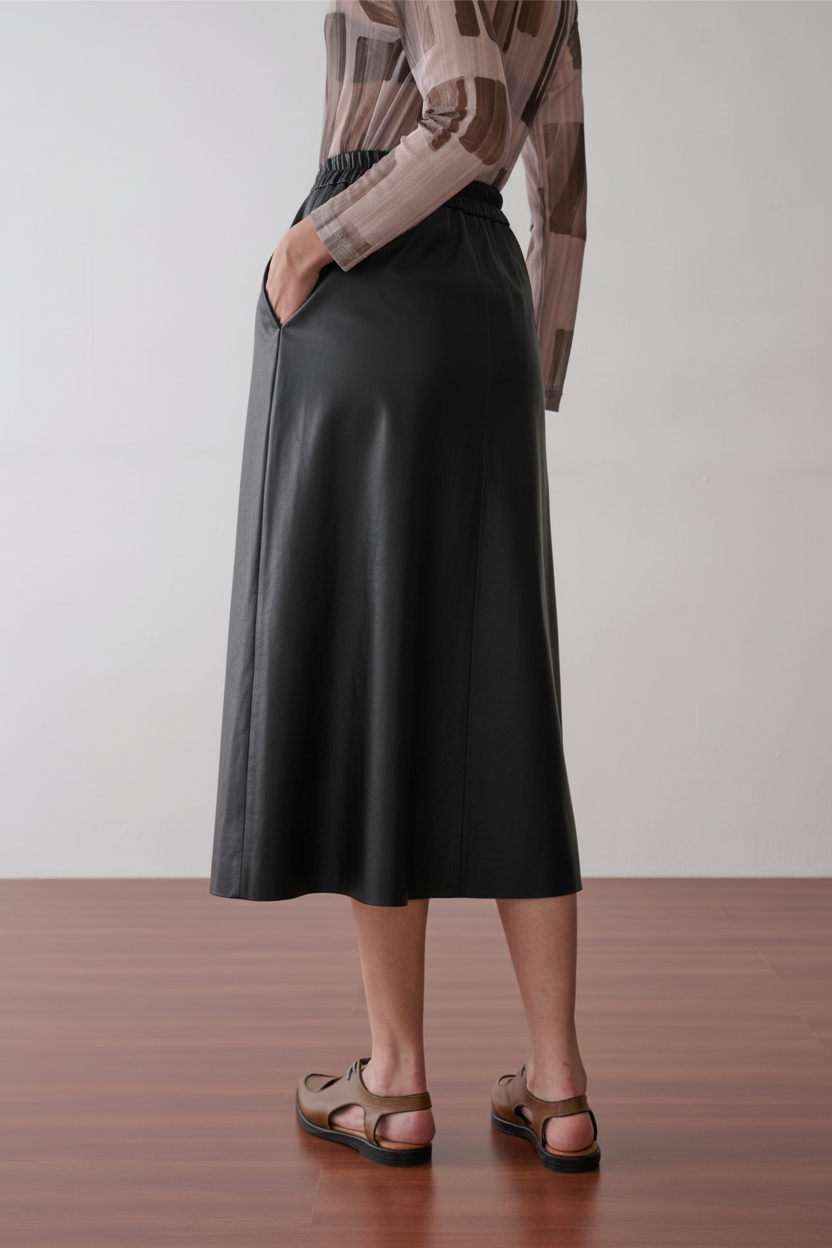 Modelo de costas usando uma saia midi preta de acabamento acetinado com cós elástico e bolsos laterais, combinada com blusa de tule estampada e sapatos marrons de bico fino.