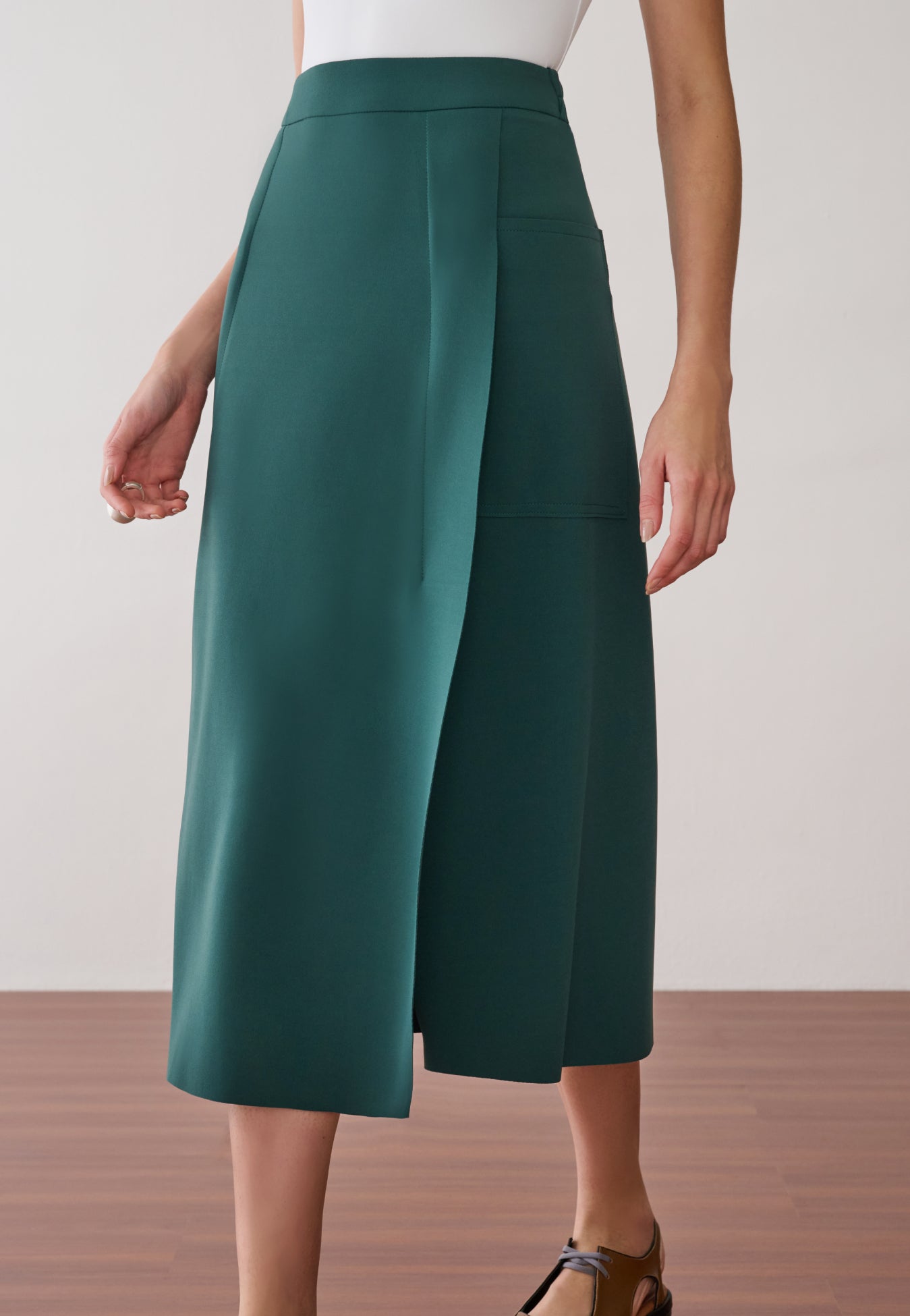 Modelo veste uma saia midi verdepetróleo com recortes laterais de acabamento acetinado e sapatos marrons de bico fino.