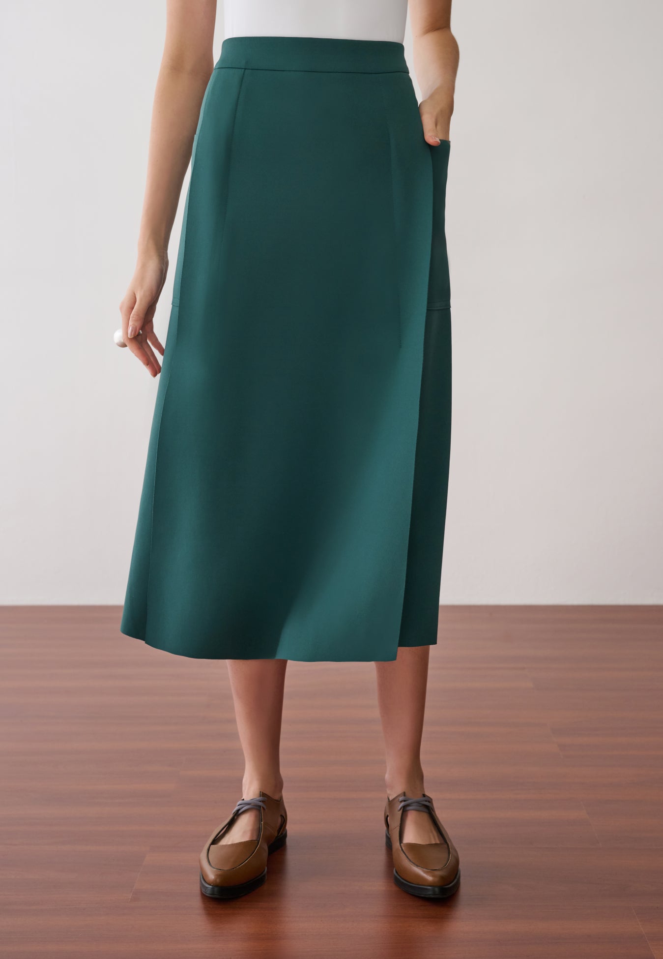 Modelo veste uma saia midi verdepetróleo com recortes laterais de acabamento acetinado e sapatos marrons de bico fino.