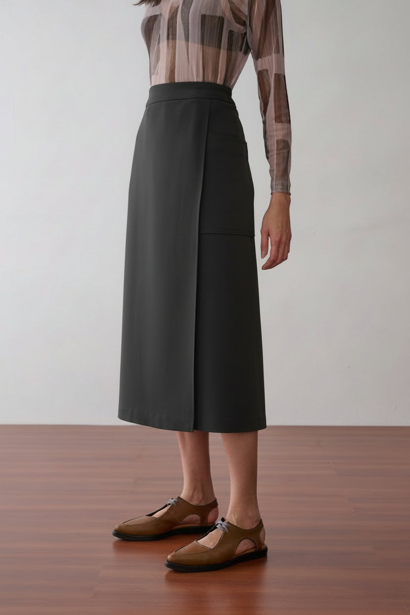 Modelo veste uma blusa de tule transparente e estampa geométrica em tons de marrom e nude, combinada com saia midi preta com recortes laterais de acabamento acetinado e sapatos marrons de bico fino.