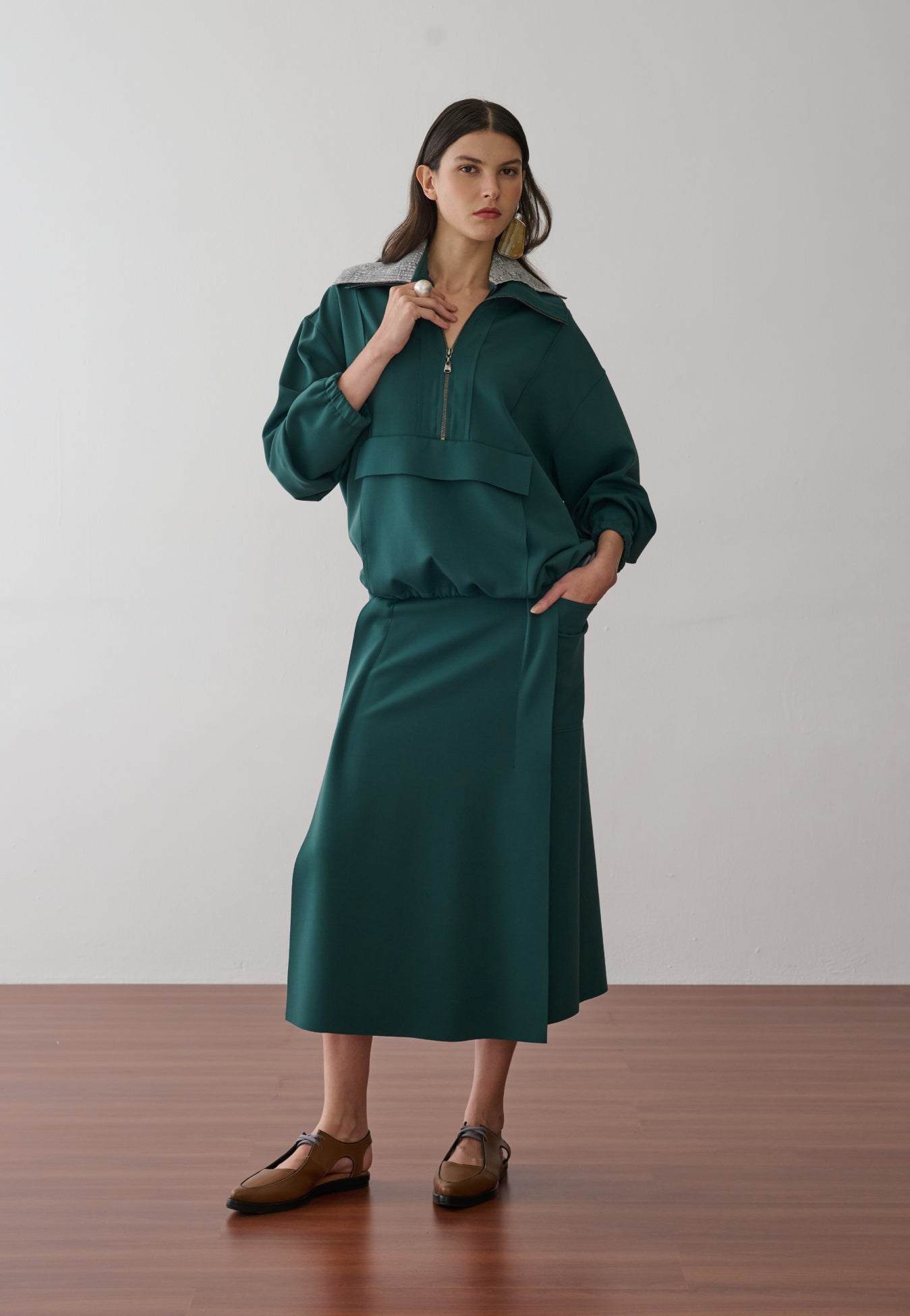 Modelo veste uma jaqueta de gola ampla e fecho em zíper na cor verde-petróleo, combinada com uma saia midi com recortes laterais de acabamento acetinado de mesma cor e sapatos marrons de bico fino.