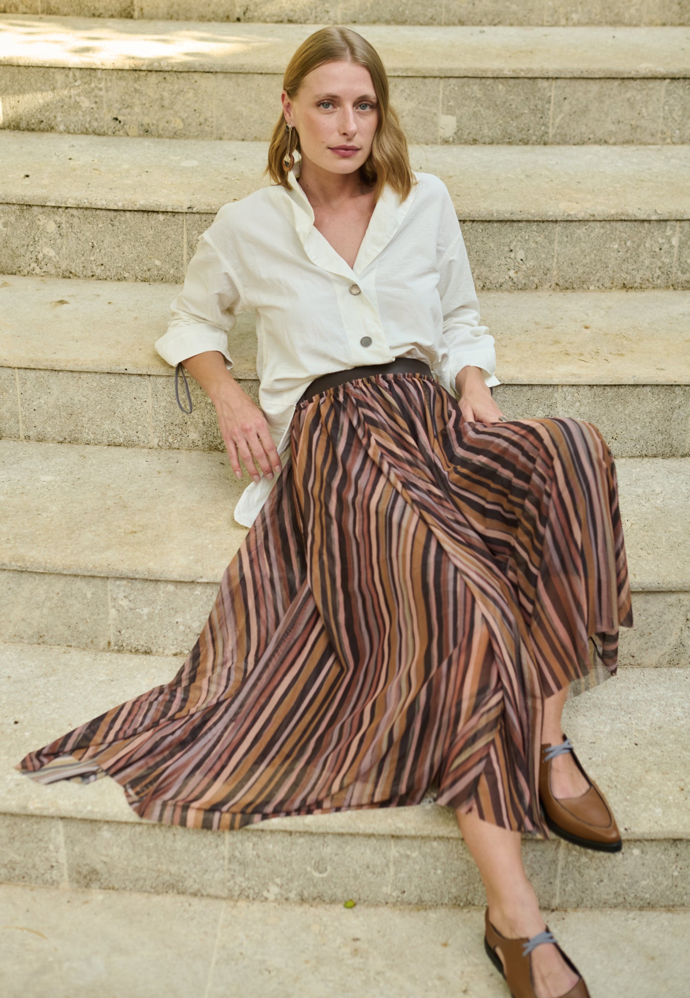 Modelo veste uma camisa off-white de mangas longas com punhos ajustáveis por cordão e reguladores metálicos, gola em "V" e botões metalizados, combinada com uma saia midi plissada em estampa multicolorida e sapatos marrons de bico fino.