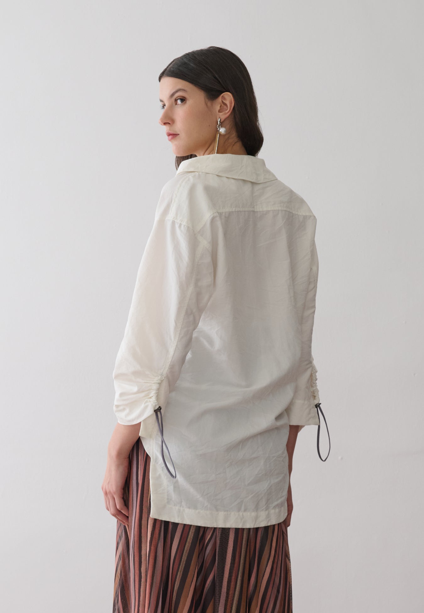 Modelo de costas veste uma camisa off-white de mangas longas com punhos ajustáveis por cordão e reguladores metálicos, combinada com uma saia midi plissada em estampa multicolorida.