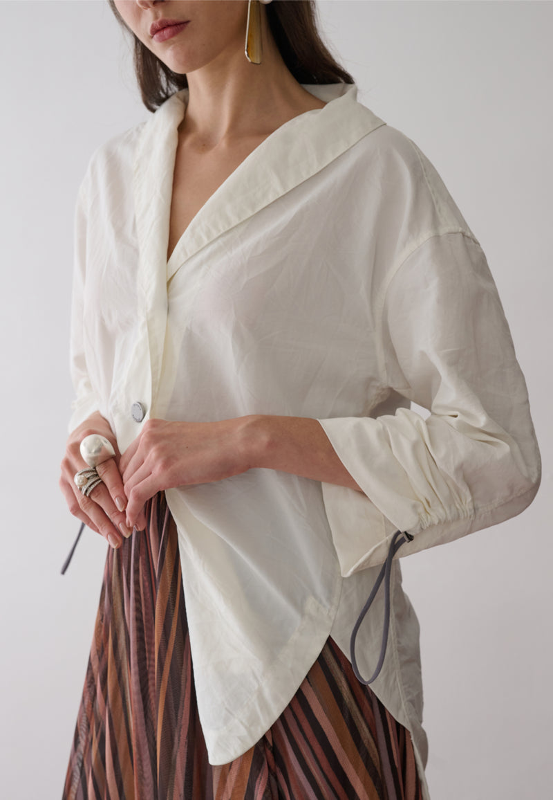 Modelo veste uma camisa off-white de mangas longas com punhos ajustáveis por cordão e reguladores metálicos, gola em "V" e botões metalizados, combinada com uma saia midi plissada em estampa multicolorida.