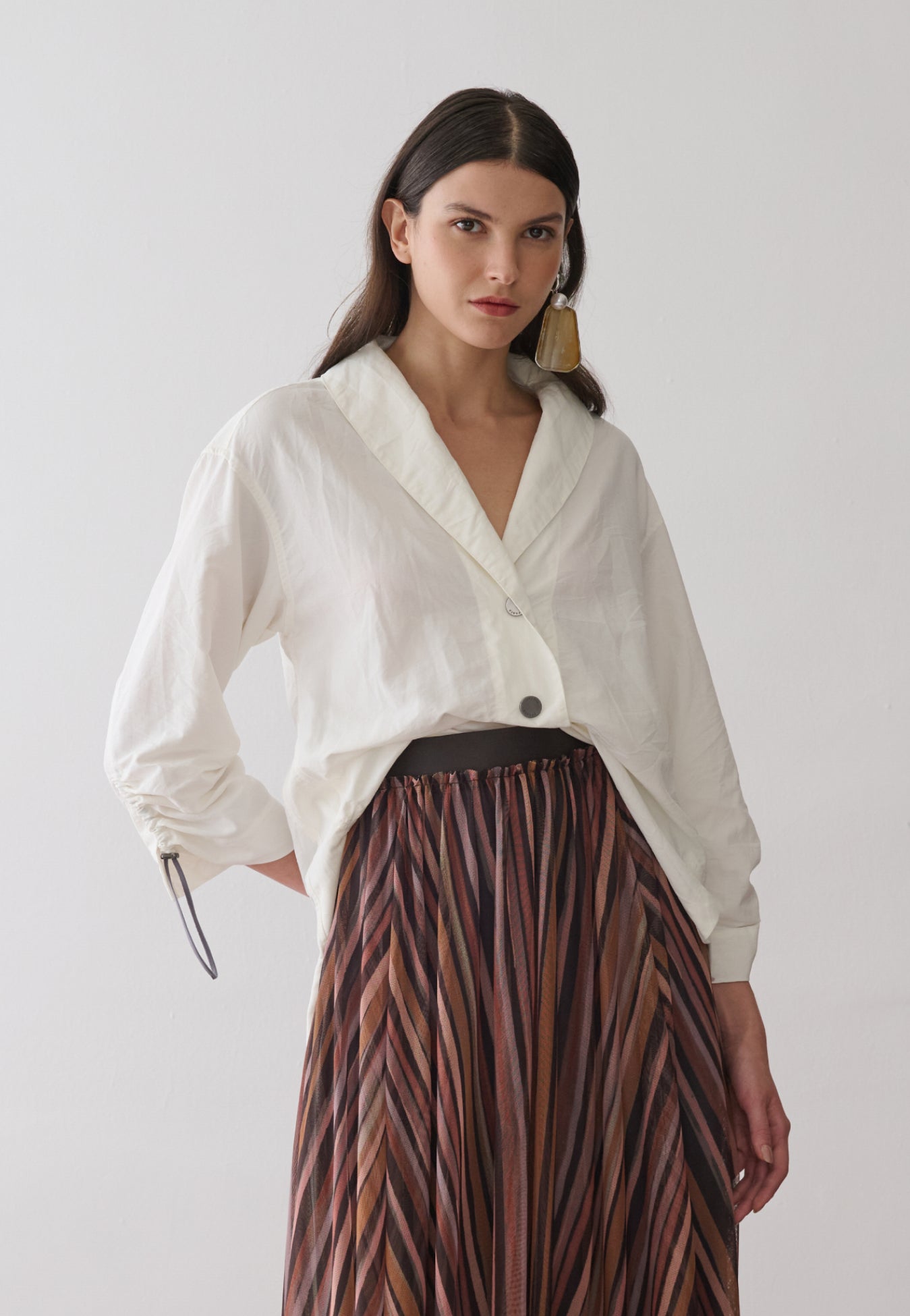 Modelo veste uma camisa off-white de mangas longas com punhos ajustáveis por cordão e reguladores metálicos, gola em "V" e botões metalizados, combinada com uma saia midi plissada em estampa multicolorida.