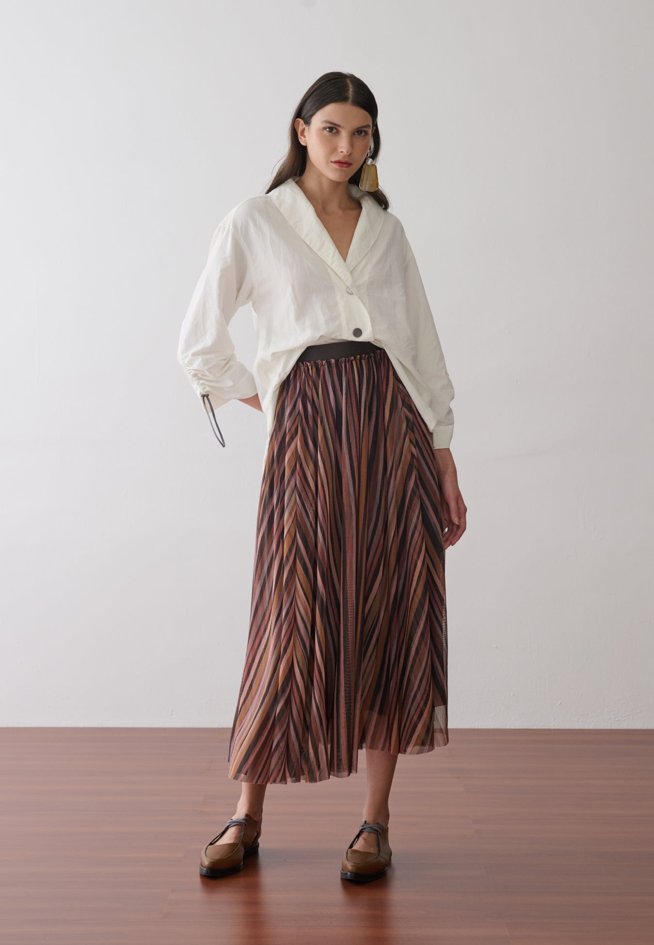 Modelo veste uma camisa off-white de mangas longas com punhos ajustáveis por cordão e reguladores metálicos, gola em "V" e botões metalizados, combinada com uma saia midi plissada em estampa multicolorida e sapatos marrons de bico fino.