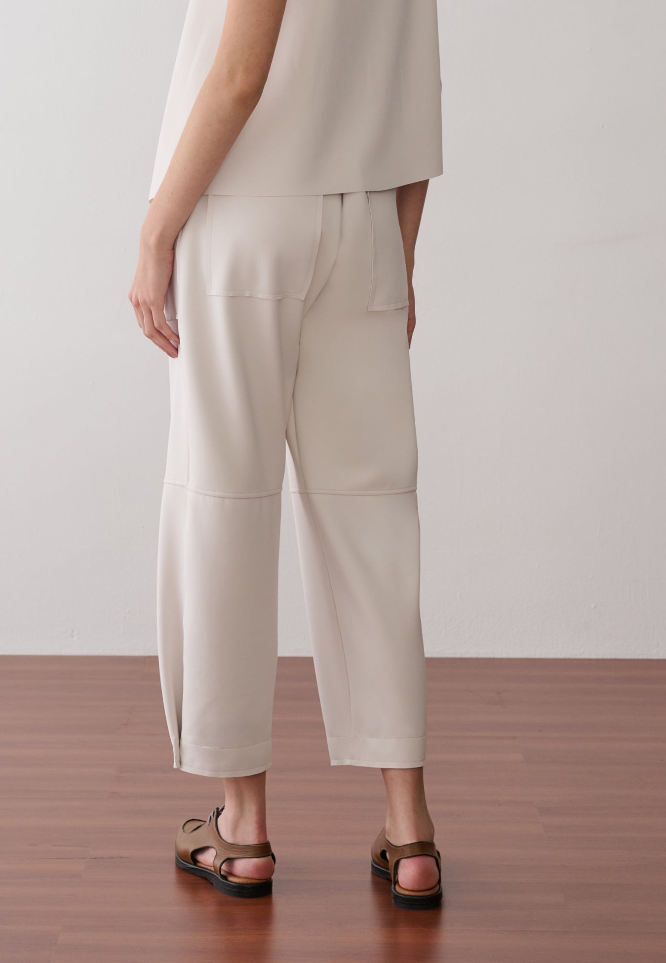 Modelo de costas veste uma calça off white de cintura alta, modelagem reta, cós elástico e bolsos amplos.