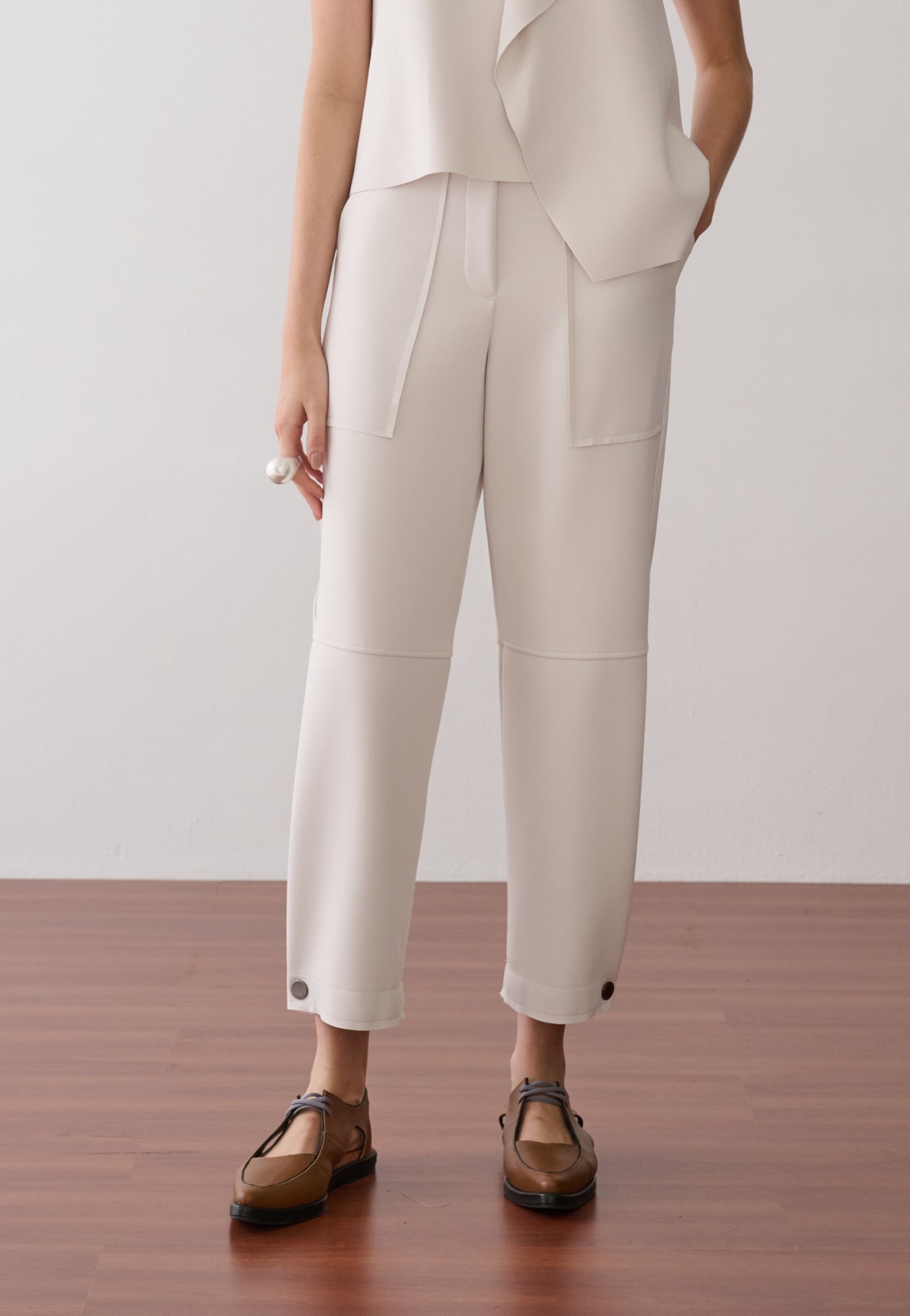 Modelo veste uma calça off white de cintura alta e modelagem reta com bolsos frontais amplos, detalhe de botões nos punhos da barra e sapatos de bico fino.