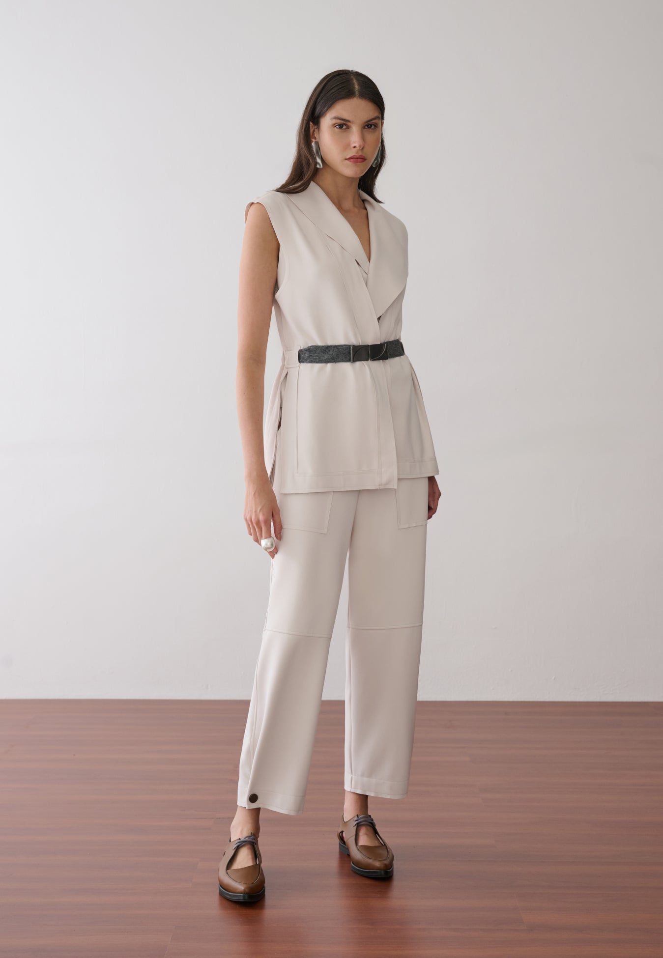 Modelo veste um conjunto off-white de colete alongado com lapelas e cinto preto sobre calça reta de bolsos amplos, finalizado com sapatos marrons de bico fino.
