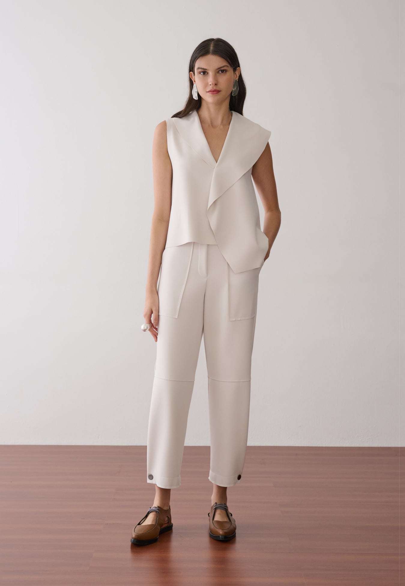 Modelo veste uma blusa off-white sem mangas com lapelas assimétricas e gola em "V", combinada com calça reta da mesma cor com bolsos amplos e sapatos marrons de bico fino.