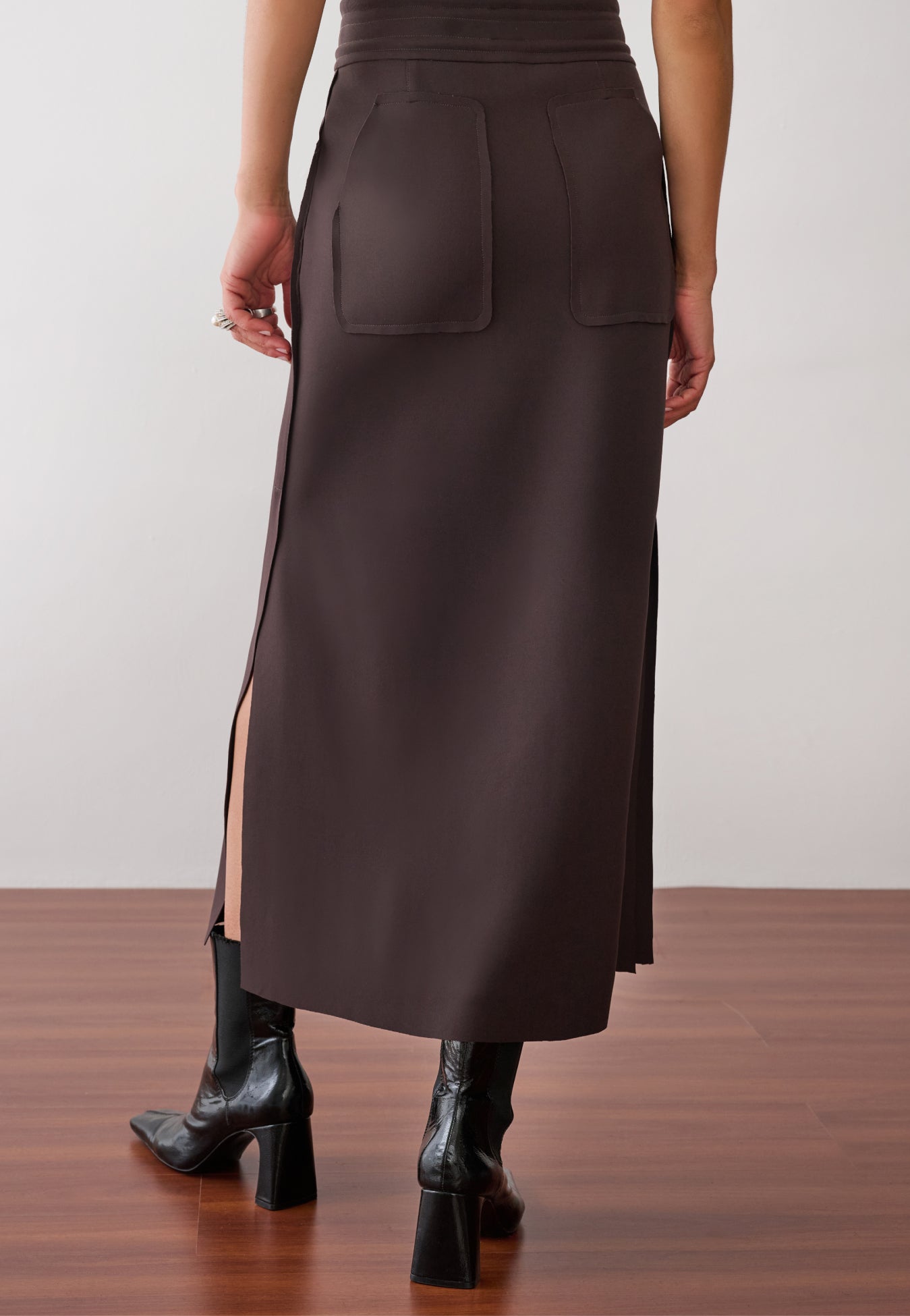 Modelo de costas usando uma saia midi marrom com fendas laterais, bolsos traseiros aplicados, cós elástico e botas pretas de verniz com bico quadrado.