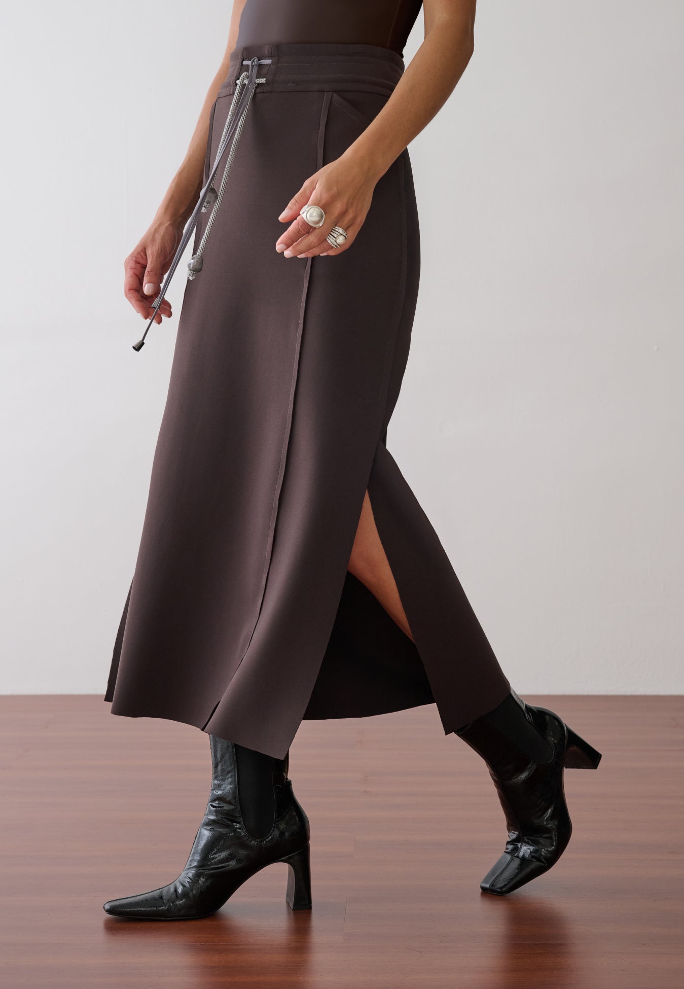 Modelo veste uma saia midi marrom que apresenta fenda lateral, recortes laterais, cordão bicolor frontal e botas pretas de verniz.