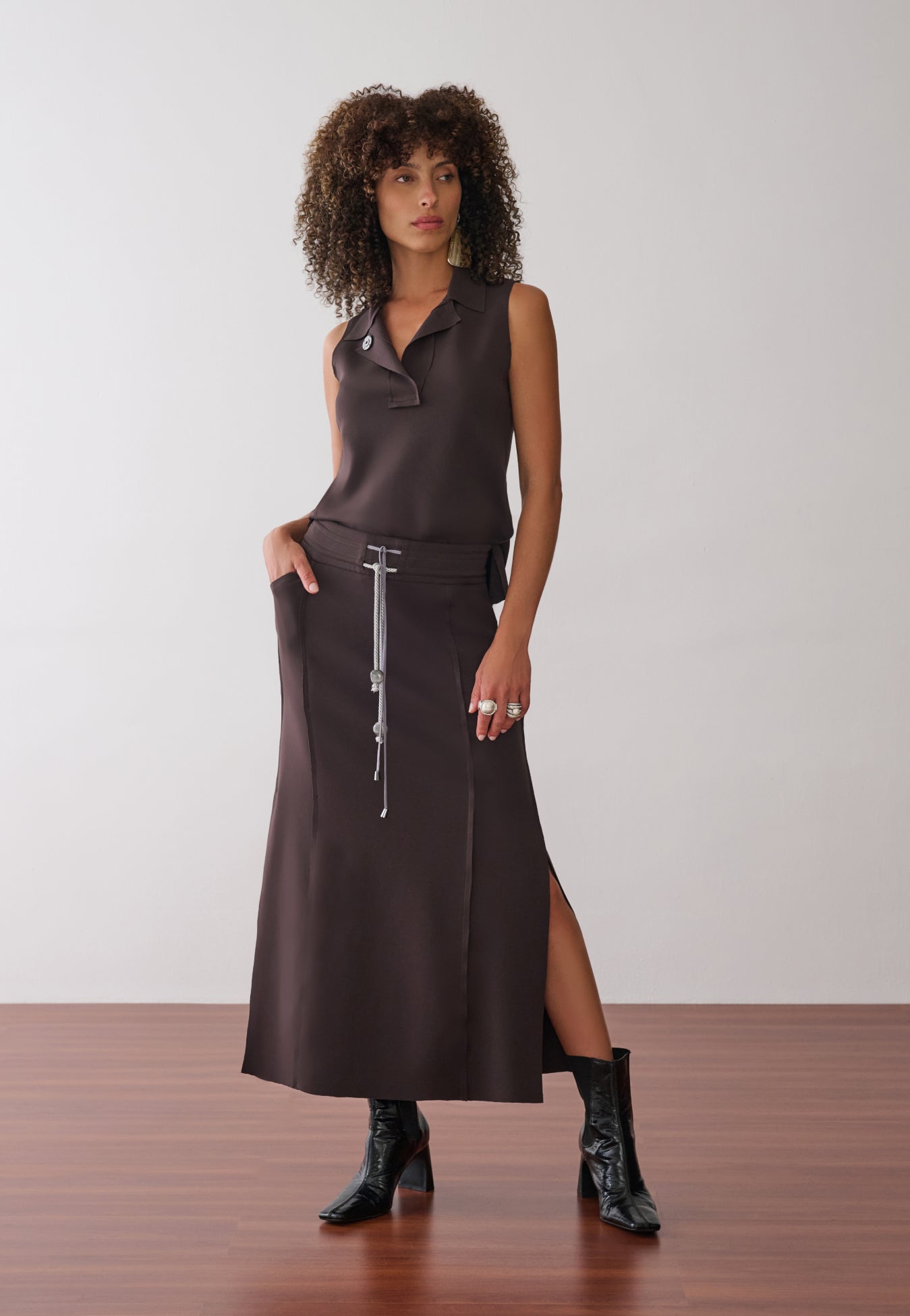 Modelo veste uma blusa marrom sem mangas com gola polo aberta, combinada com uma saia midi de mesma cor que apresenta fenda lateral, cordão bicolor com ponteiras, recortes nas laterais e botas pretas de verniz.