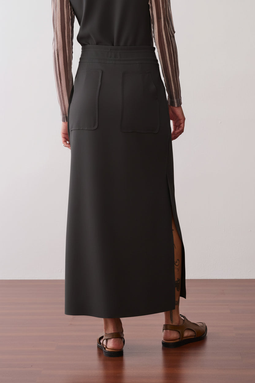 Modelo de costas usando uma saia midi preta com fendas laterais, bolsos traseiros aplicados, cós elástico e sapatos marrons.